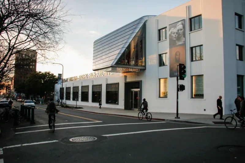 A white and glass museum building at a street corner, with bikes and a few pedestrians at the curb.