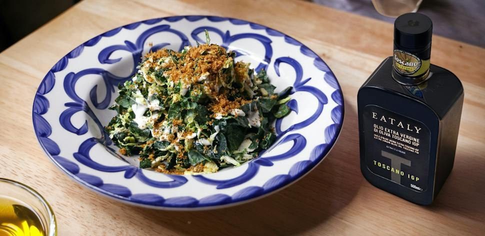 Bowl of greens with toasted breadcrumbs on a patterned blue and white plate next to a bottle of olive oil