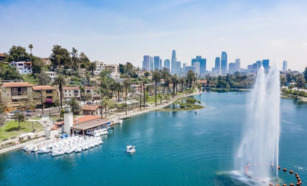 An aerial view of a lake with a tall fountain, swan boats at a dock, and downtown towers behind.