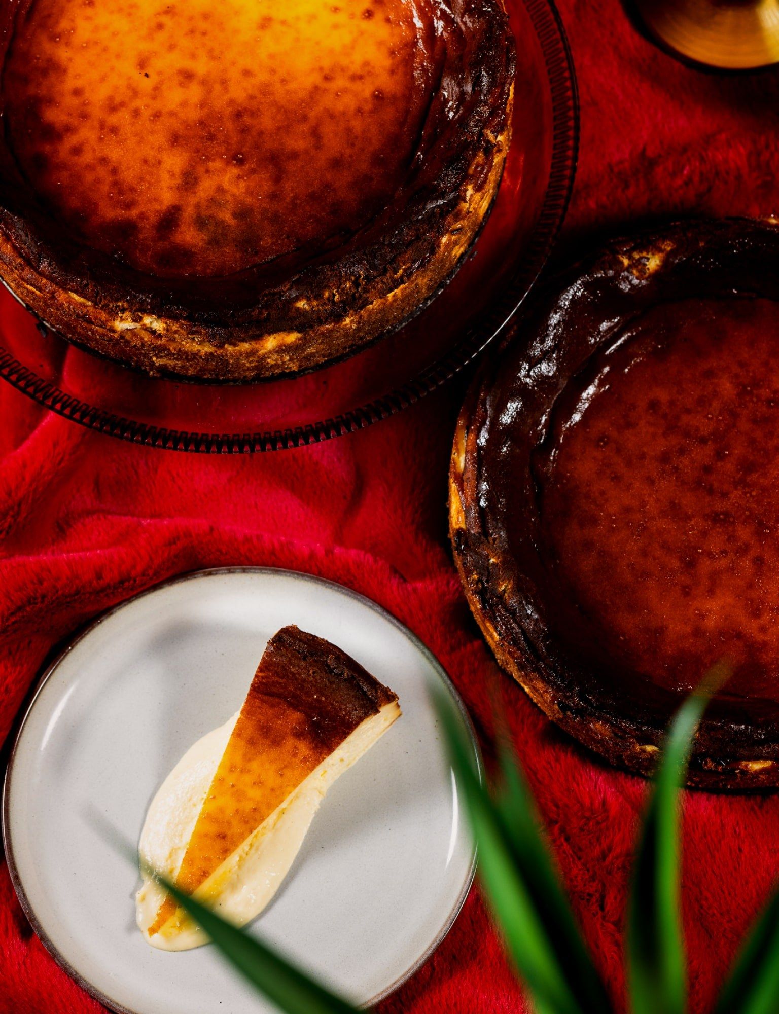 Slices of basque cheesecake on a white plate beside whole cheesecakes on a red cloth