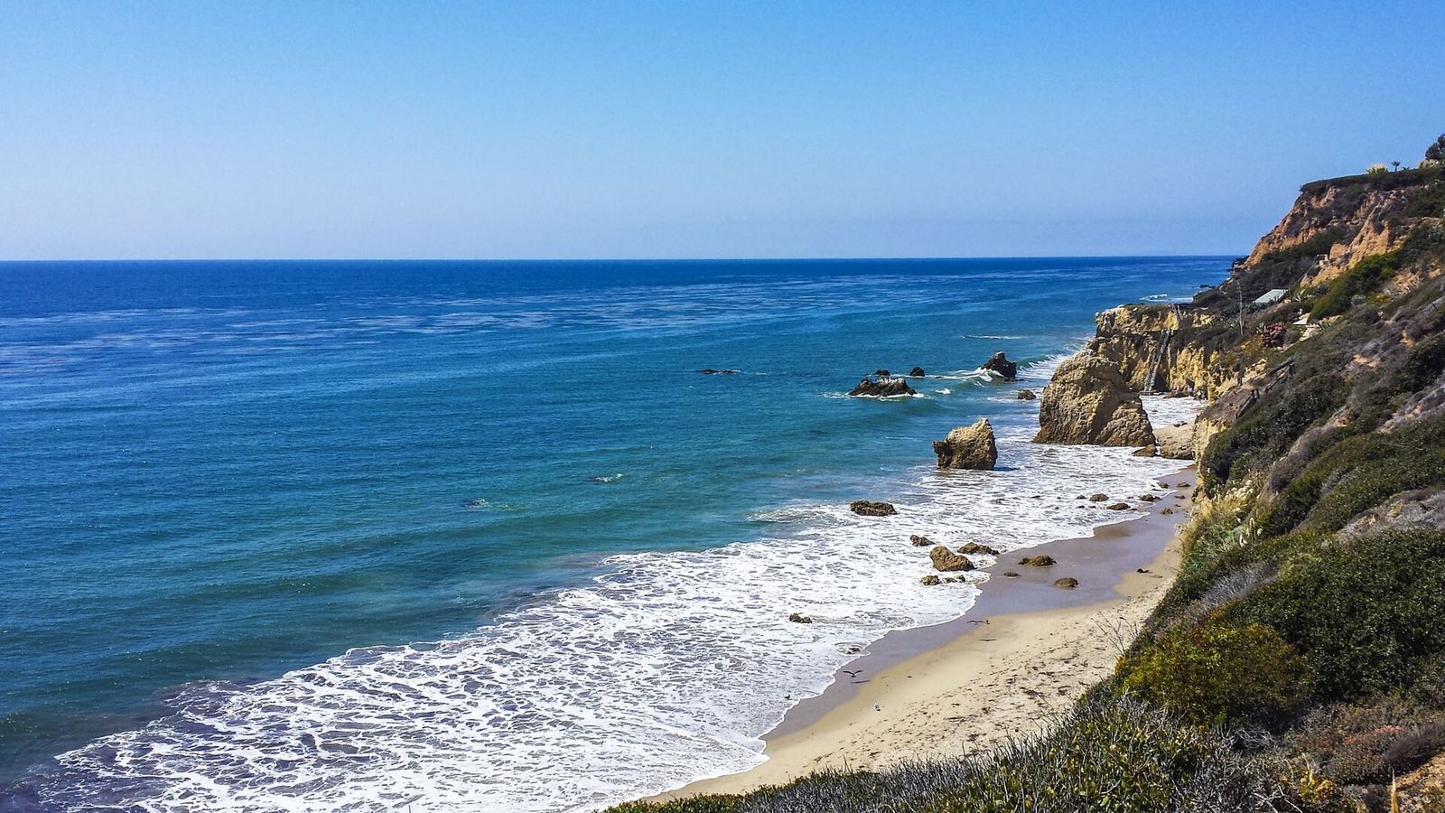 A sandy cove with waves washing over rocks and a bluff on the right under a clear blue sky.