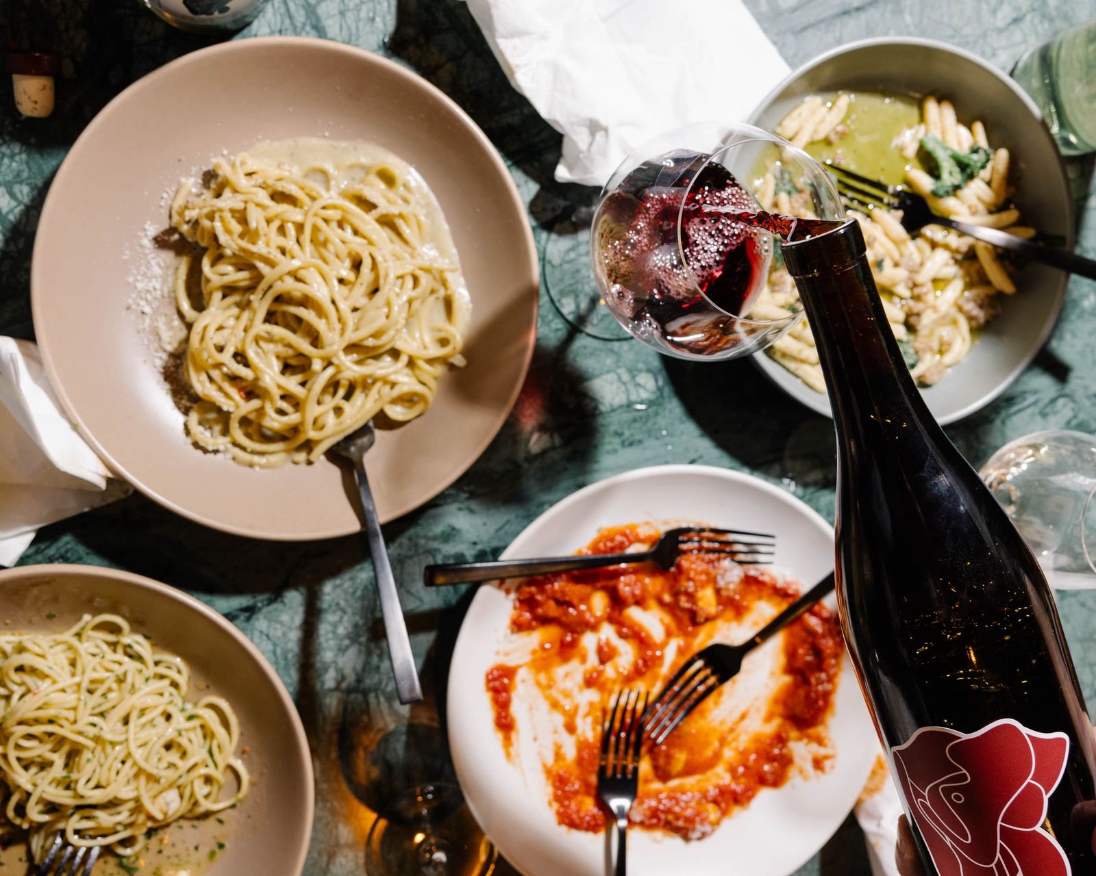 Plates of spaghetti and a tomato pasta on a green marble table with a bottle of red wine being poured