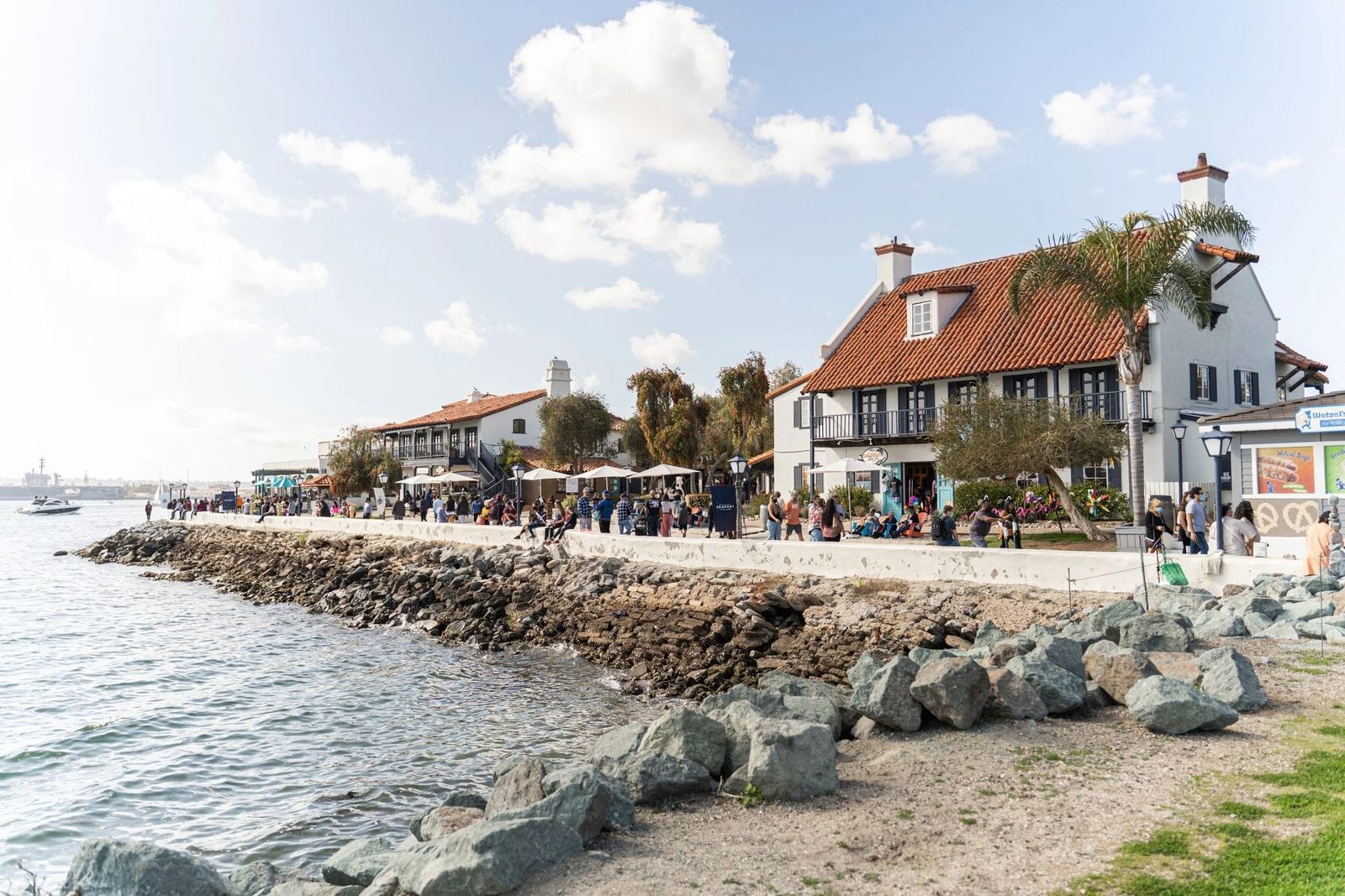 A rocky shoreline and waterfront walkway leading to shingled shops and a lighthouse-style tower under a cloudy sky.