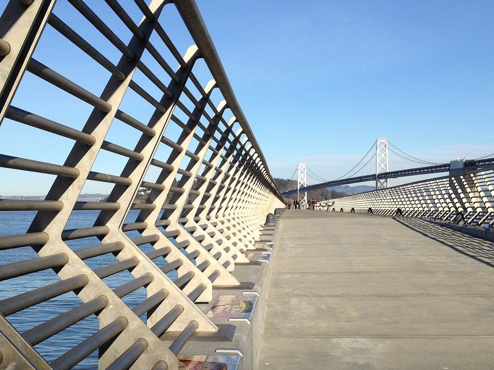 A long pier deck with angled steel railings leading out toward the Bay Bridge on a clear day.