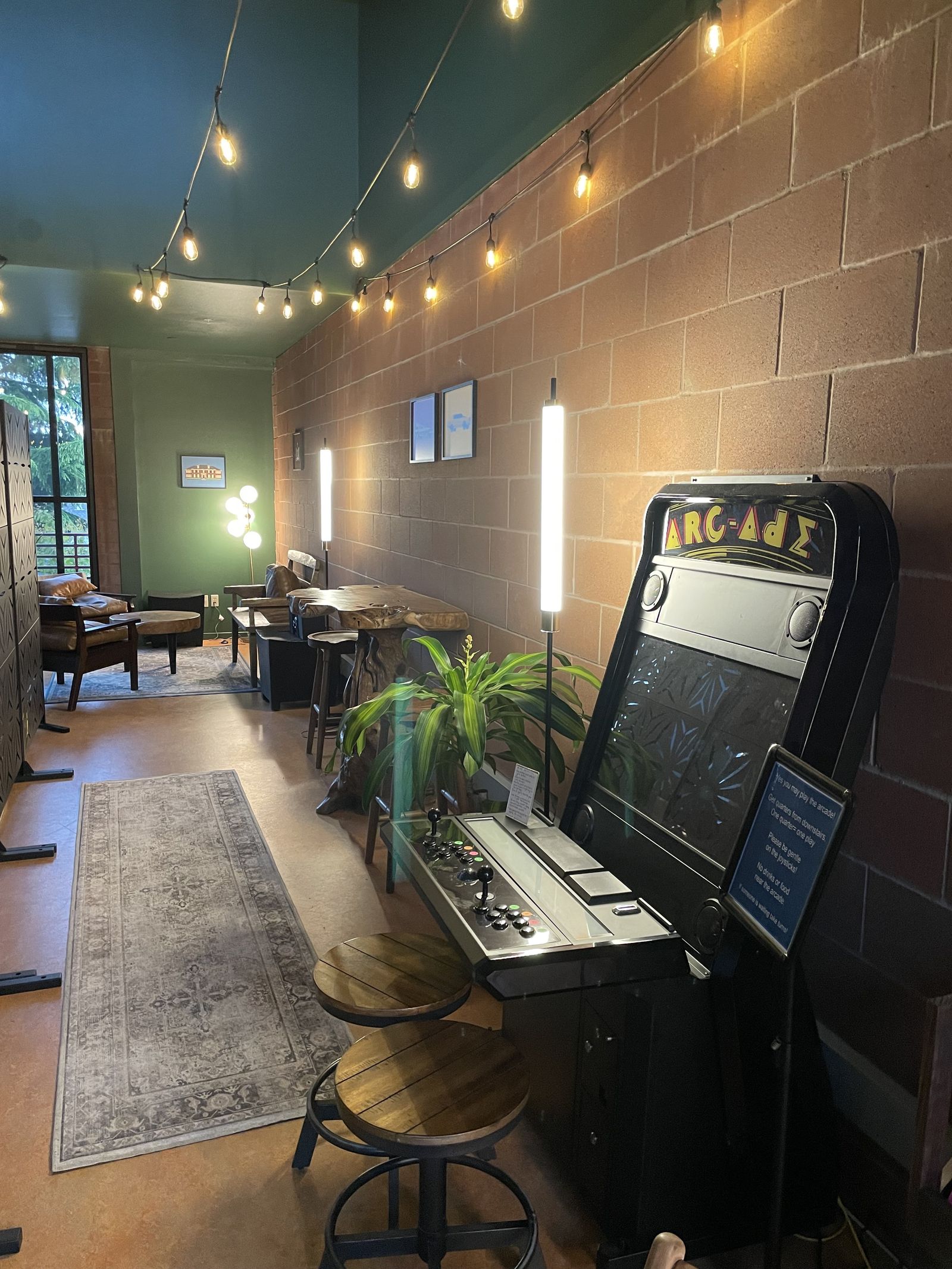 A narrow room with a brick wall, string lights, an arcade cabinet and a few seats along the side.