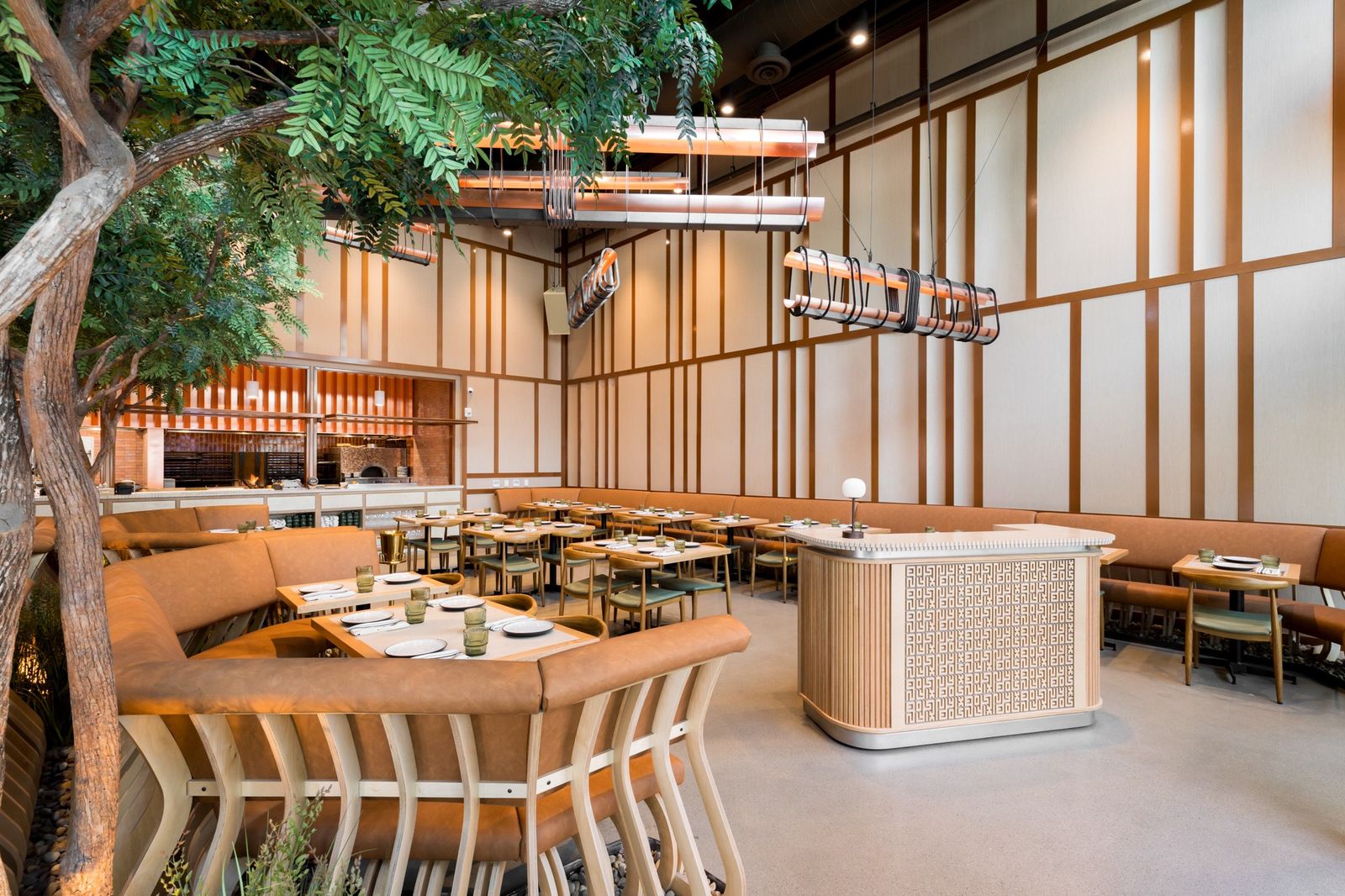 A bright restaurant interior with tables and curved wooden booths under tall trees and slatted wood walls