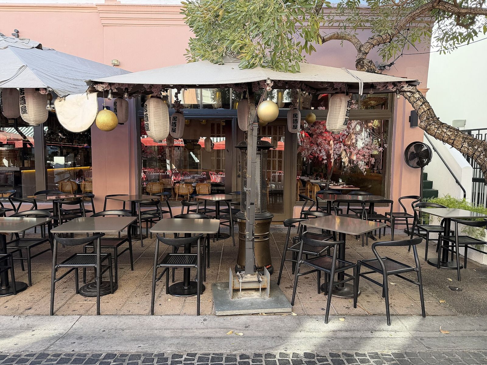 Sidewalk cafe tables under umbrellas with paper lanterns in front of a pink building