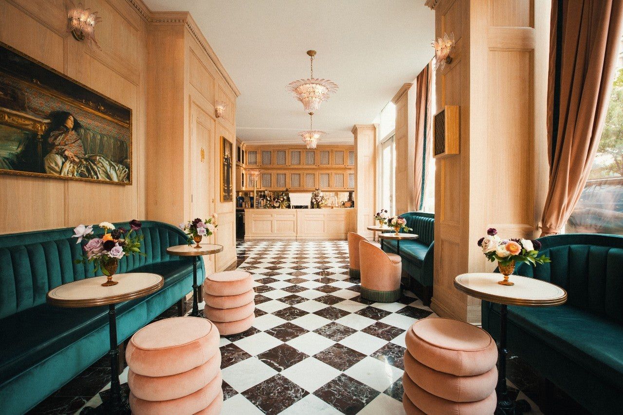A cafe with green velvet banquettes, a checkered floor, pink poufs and tall windows with curtains.