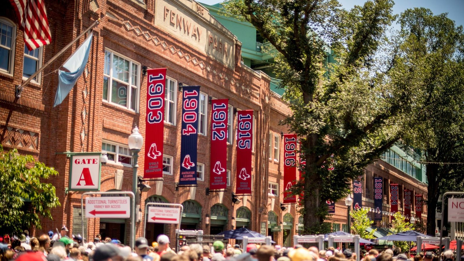 The brick exterior of Fenway Park at Gate A with red banners and a crowd outside