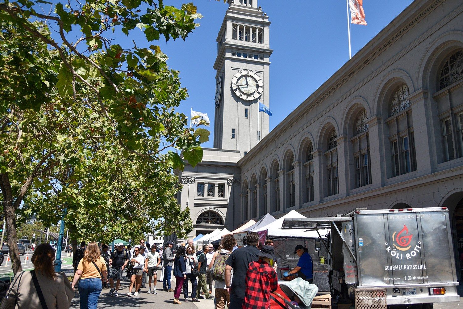 The clock tower of the Ferry Building above a crowd walking between market stalls under trees.