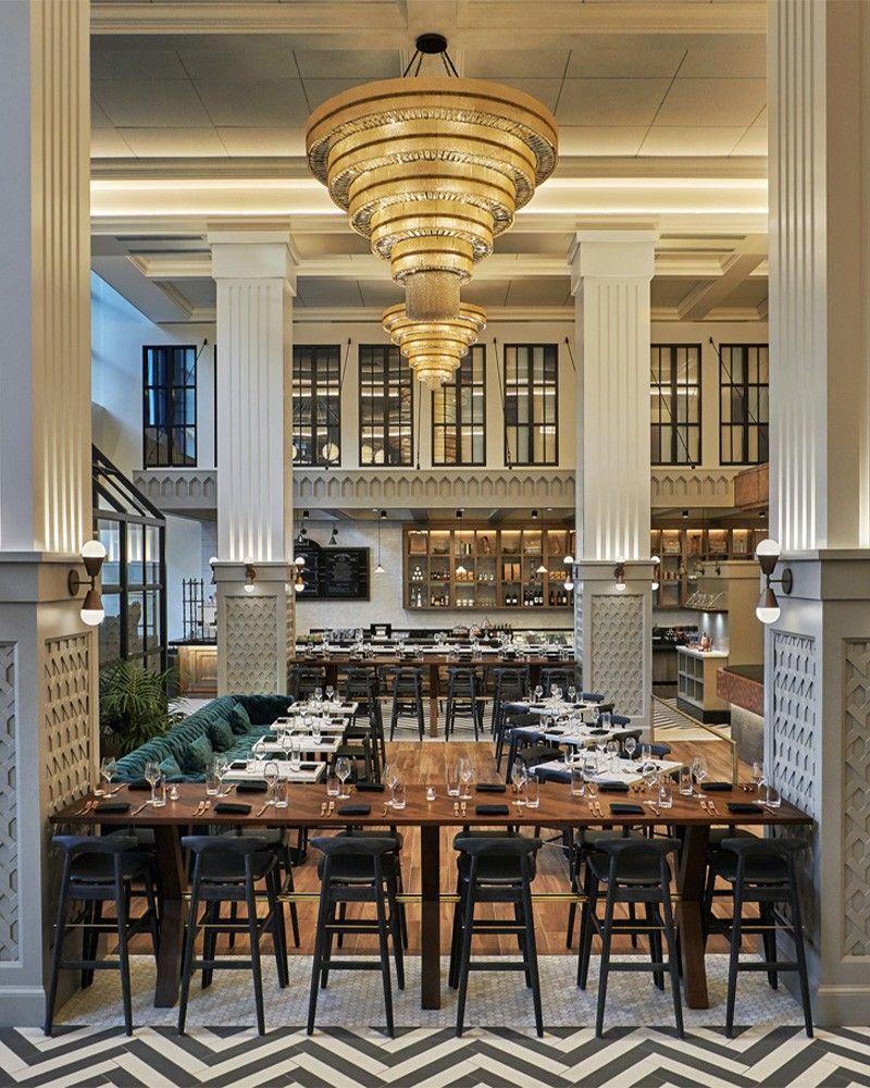 A tall dining room with columns, a large chandelier and rows of tables set with chairs under a coffered ceiling.