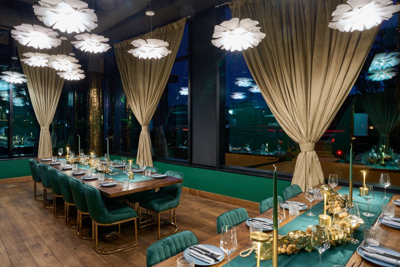 A long dining table with green velvet chairs and gold decor in a dining room with tall draped windows