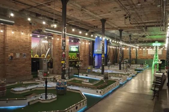 An indoor mini-golf course in a brick warehouse space with string lights, black columns and lit green fairways.