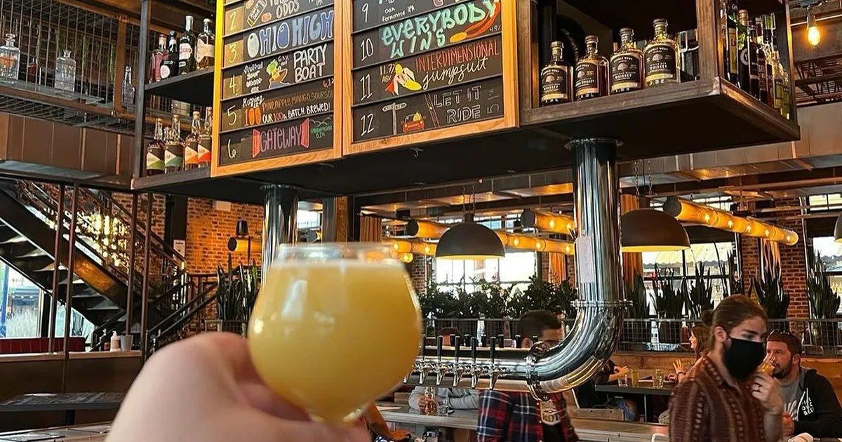 A hand holding a glass of hazy beer in front of a tiered bar with shelves of bottles and taps.