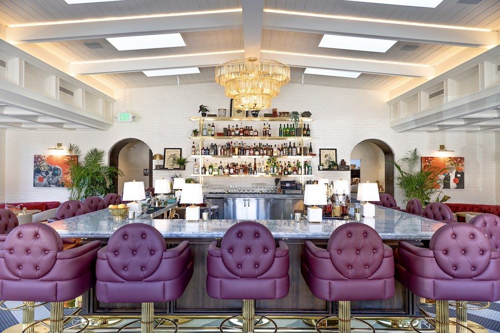 A bright bar room with a lit back bar, purple tufted stools and a chandelier under a coffered ceiling.