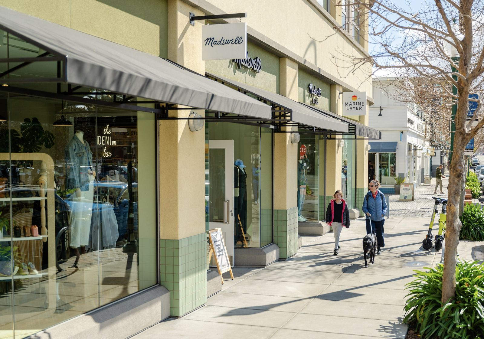 A shaded sidewalk of shops with awnings and a few pedestrians under bare trees.