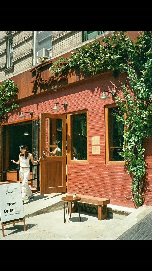 A red-brick storefront with wooden doors, green vines and a sandwich board, on a sunny corner.