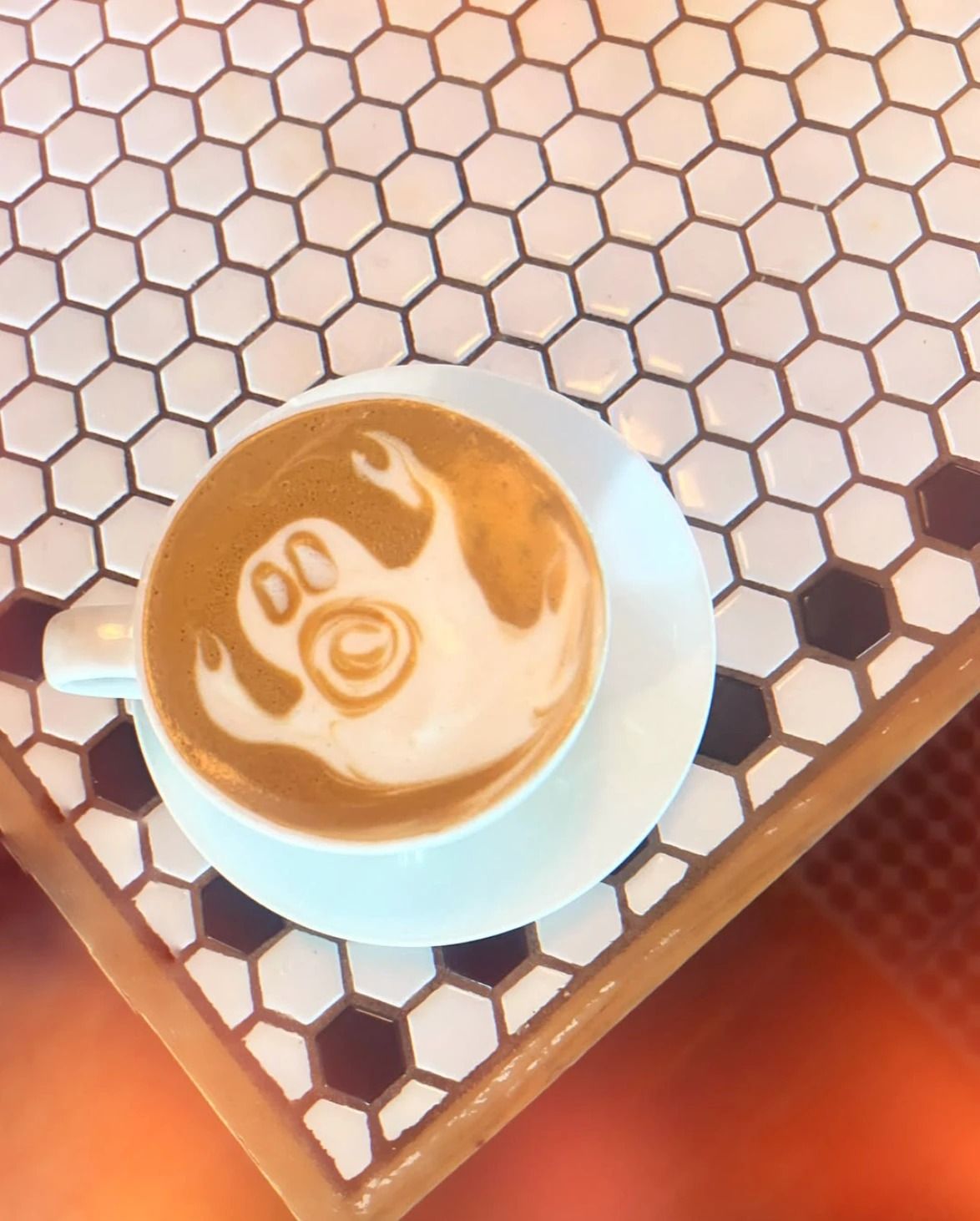 A latte with a ghost drawn in the foam, on a saucer on a white hexagon-tiled table.