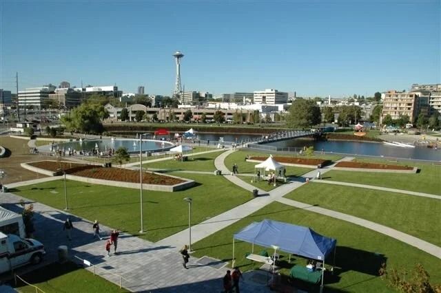 A park with lawns, curving paths and a footbridge over water, with the Space Needle and city buildings in the distance.