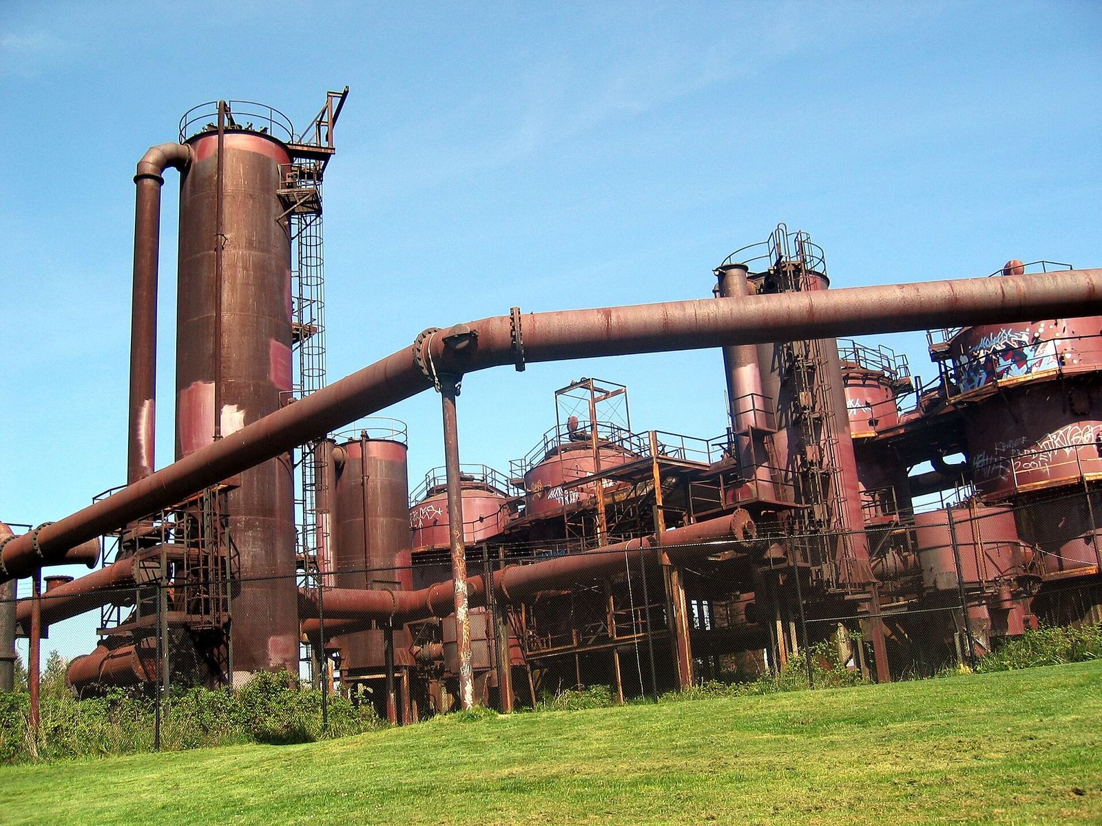 Rusted industrial towers and pipes rising above a green lawn under a blue sky.