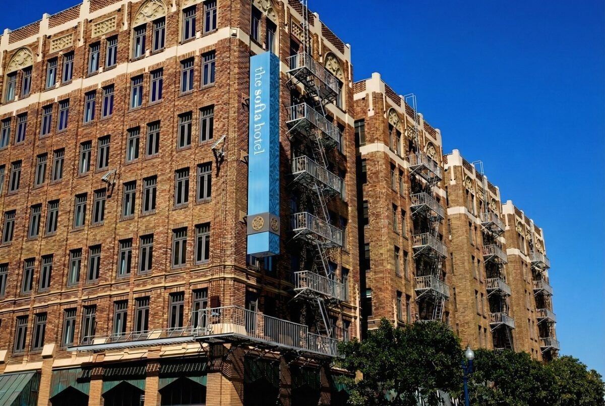 A tall brick building with fire escapes and a blue vertical sign, seen from the street under a blue sky.