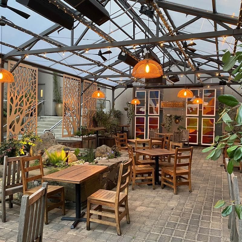 A covered patio with wooden tables and chairs, orange pendant lamps and screens of plants.