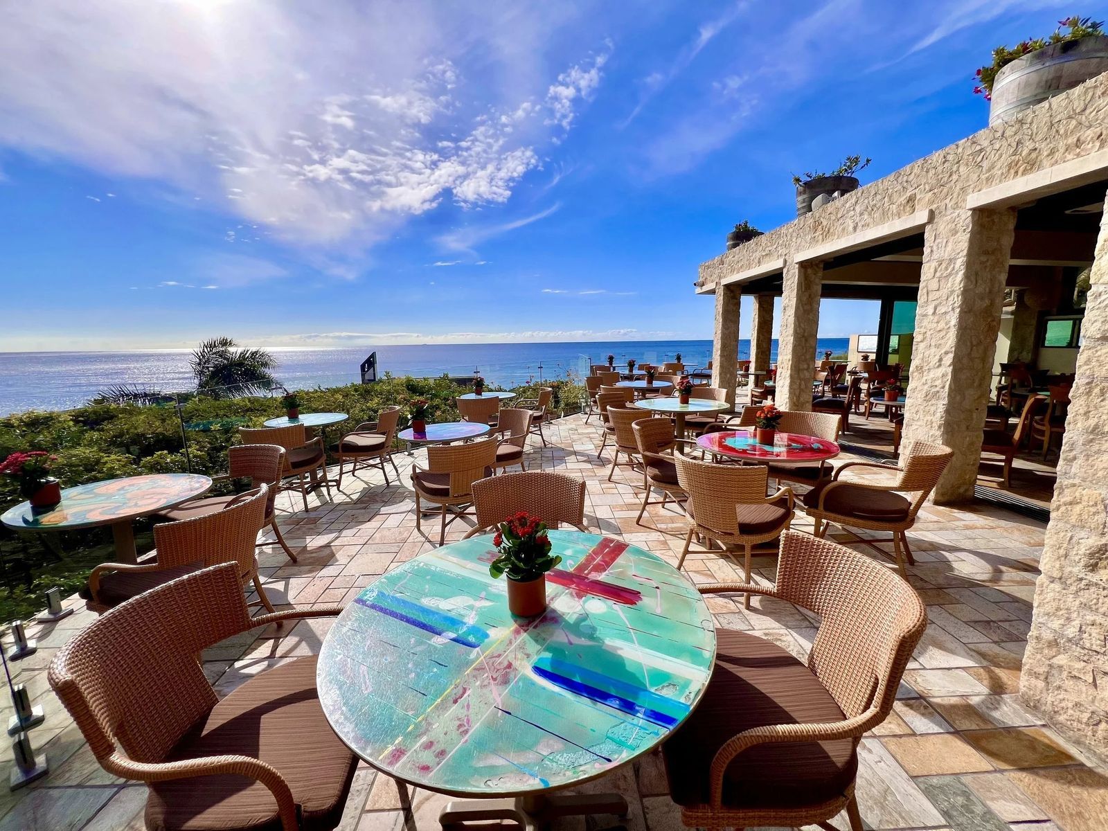 An ocean-view patio with rattan chairs, round tables and a colorful mosaic table in the foreground.