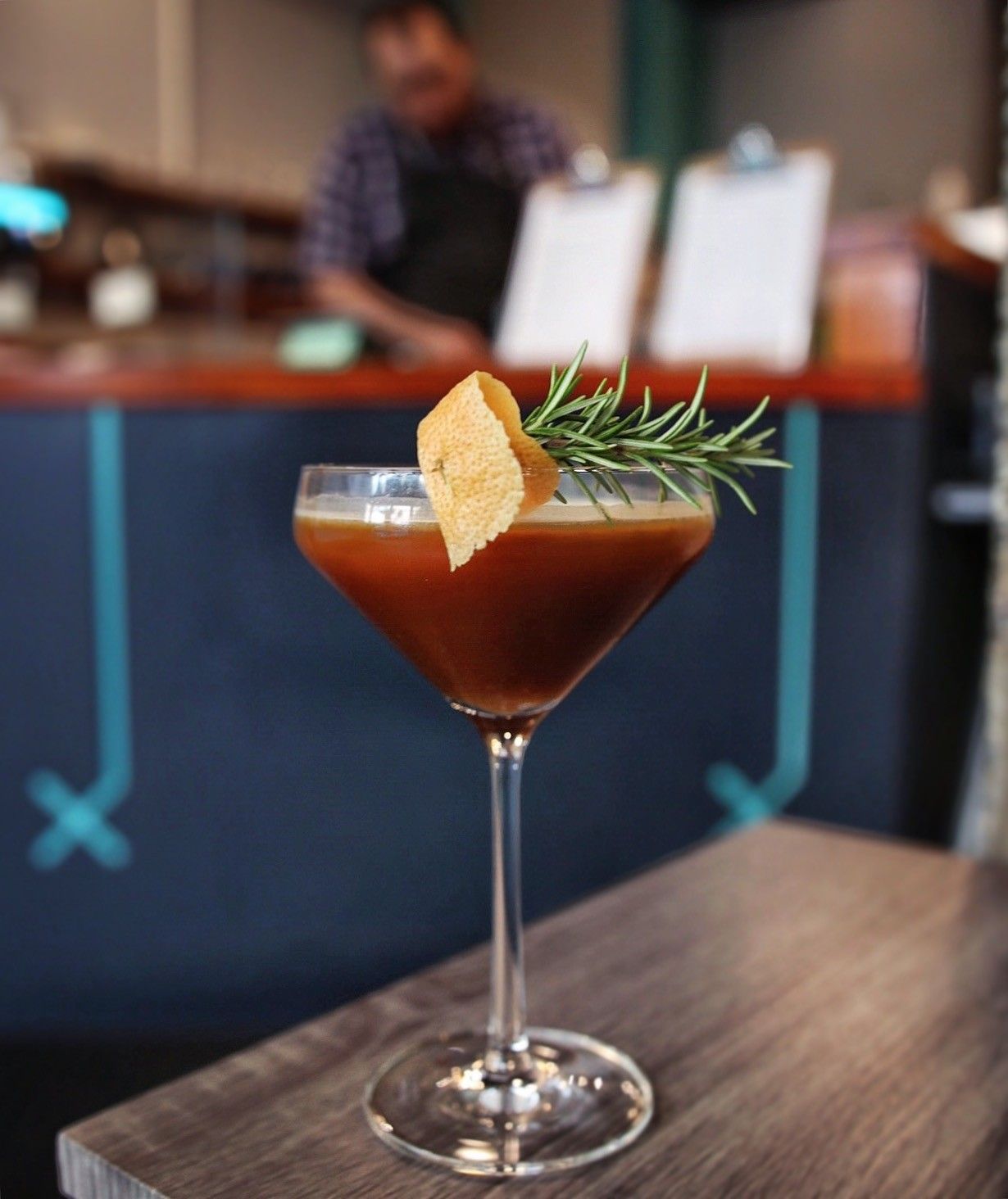 An espresso martini garnished with rosemary and a wedge of shortbread on a wooden bar, with a barista blurred behind it.