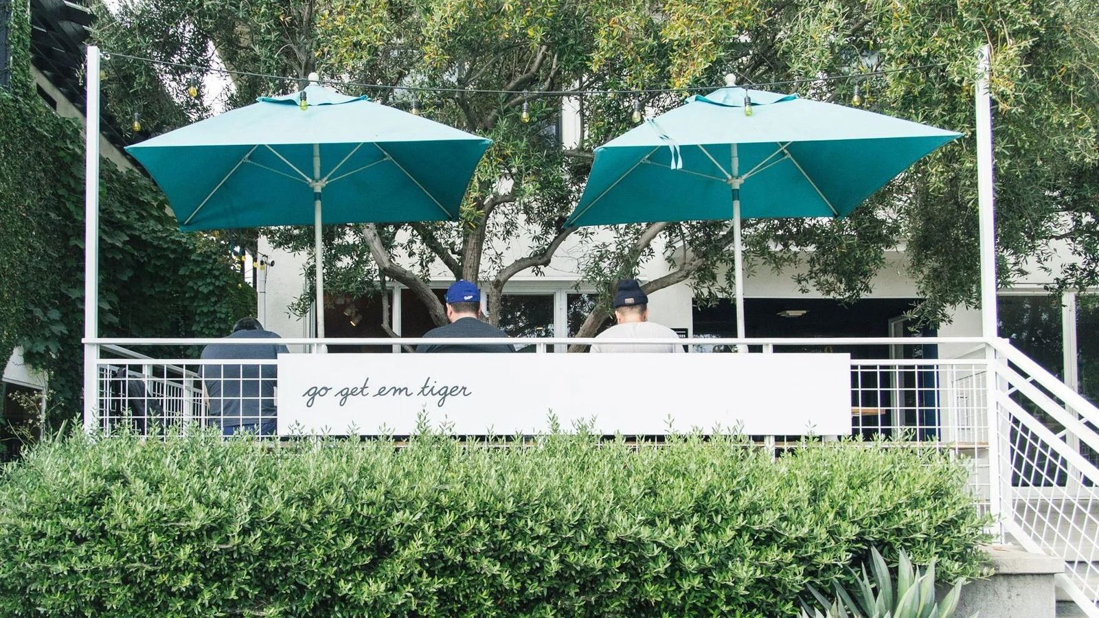A patio with teal umbrellas and tables behind a low white wall and hedge.