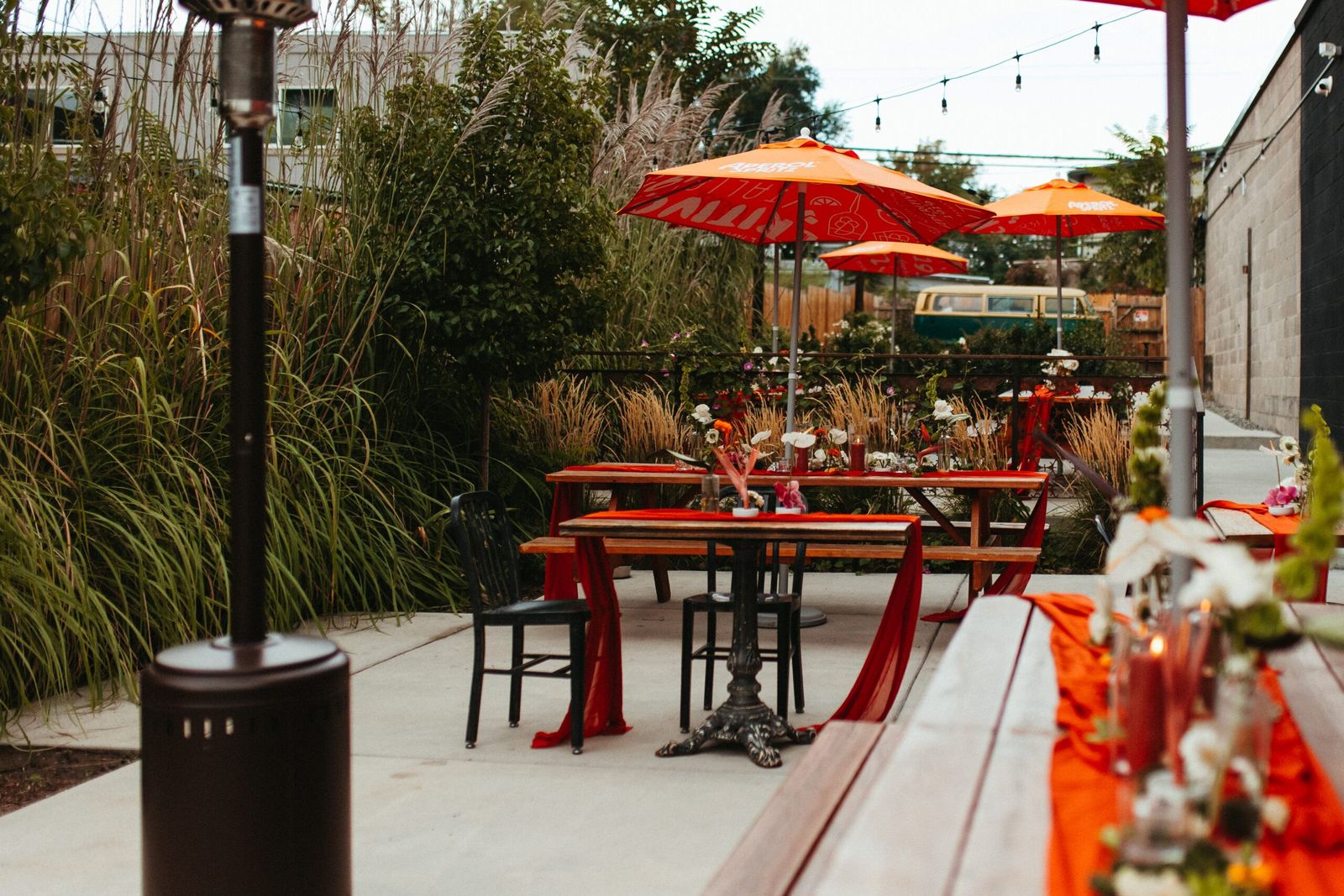 A patio with red umbrellas, tables and string lights beside tall grasses and a heater.