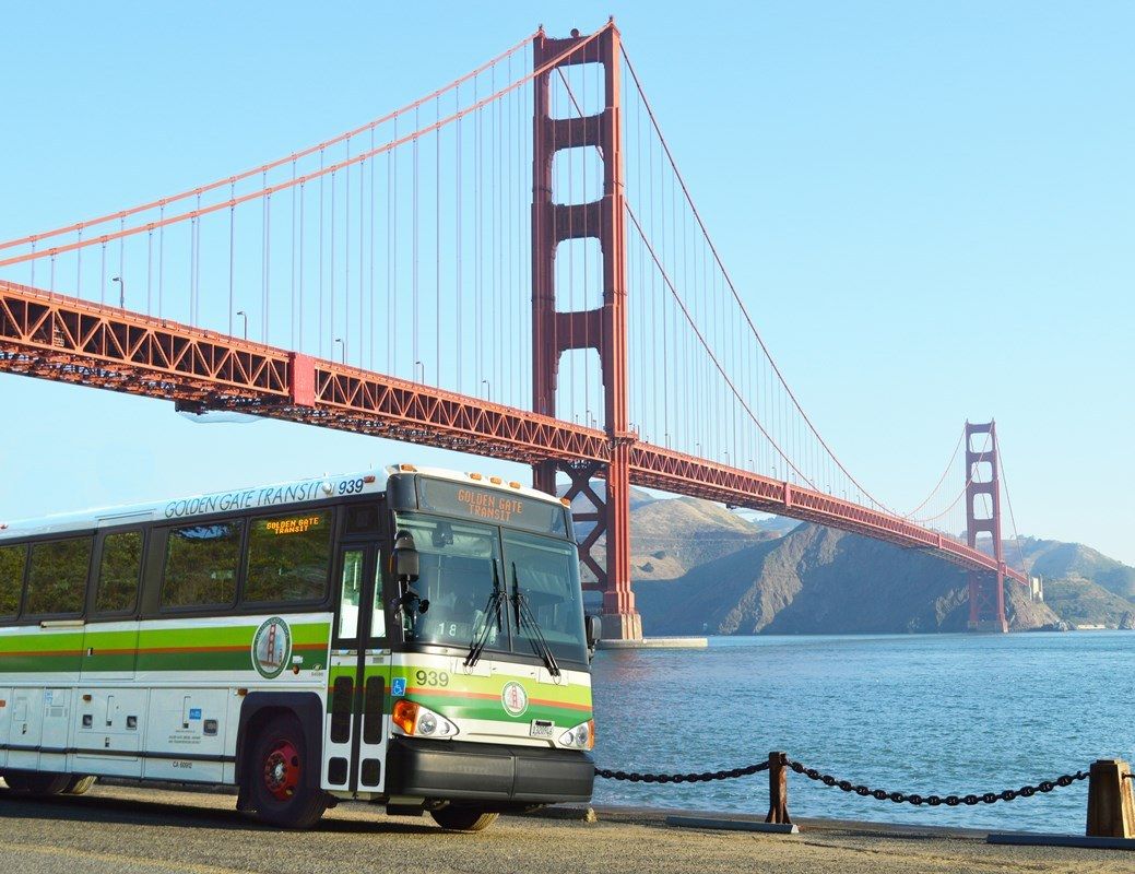 The red suspension bridge towers seen over the water with a bus parked on the shoreline in front.