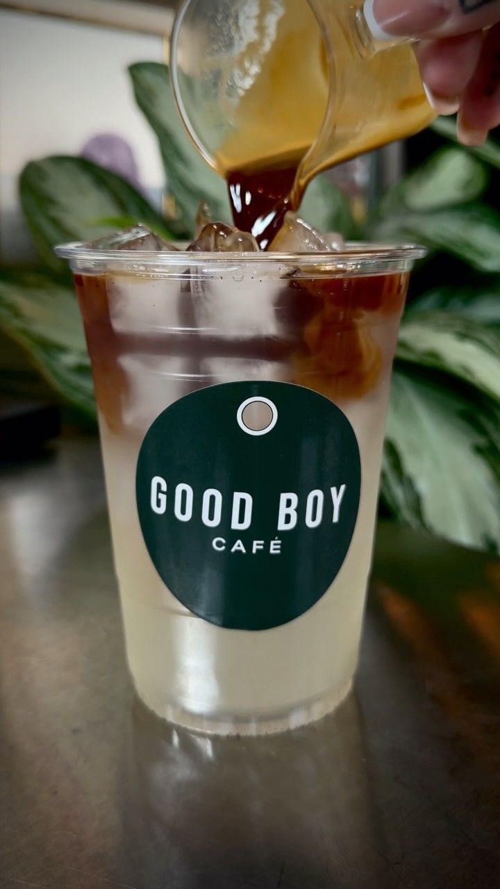 An iced coffee being poured into a clear cup printed with the Good Boy Cafe logo, with a leafy plant behind.