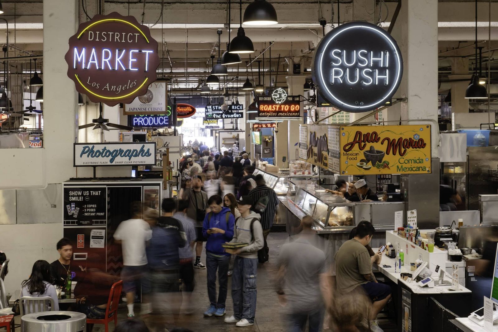 A busy market hall with food stalls under hanging lamps and neon signs, with a Market sign in the middle.
