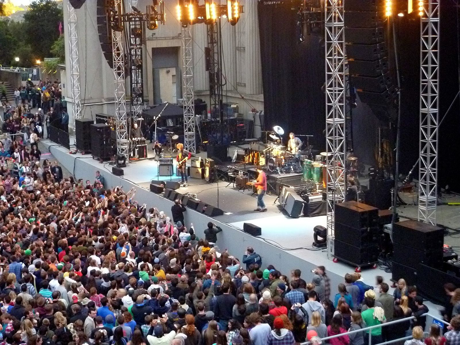 A packed outdoor amphitheater crowd facing a lit stage with a classical backdrop at dusk.