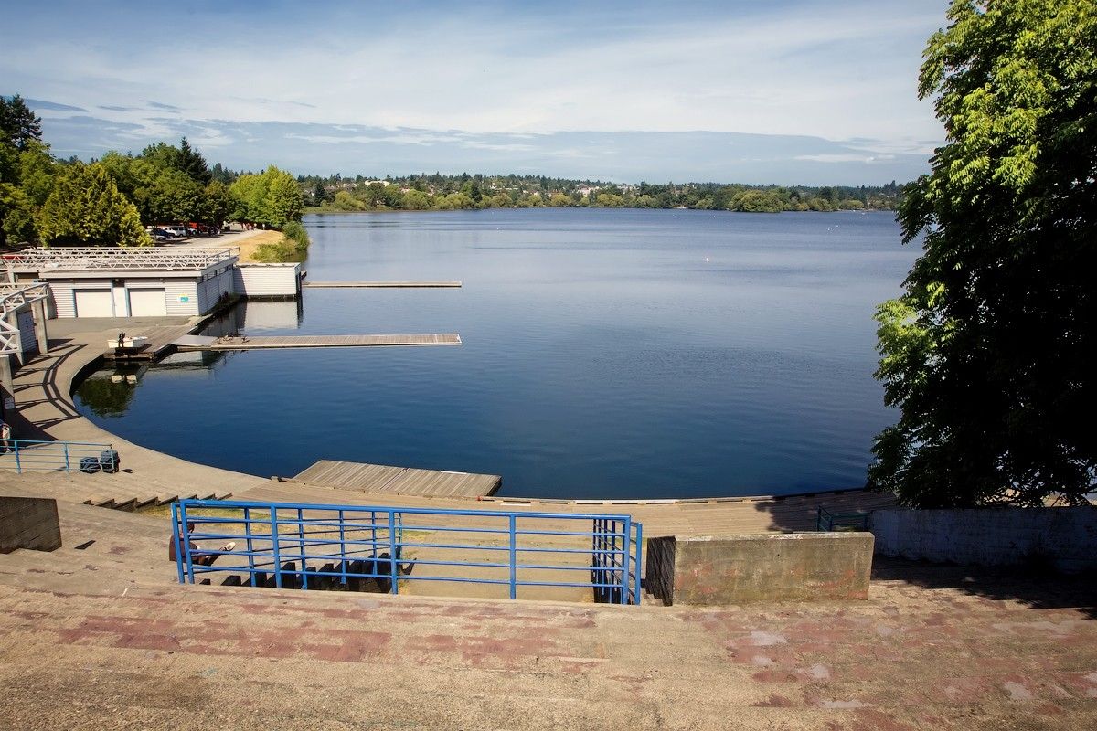 A wooden dock and viewing platform at the edge of a lake, with tree-lined shores in the distance under a partly cloudy sky.