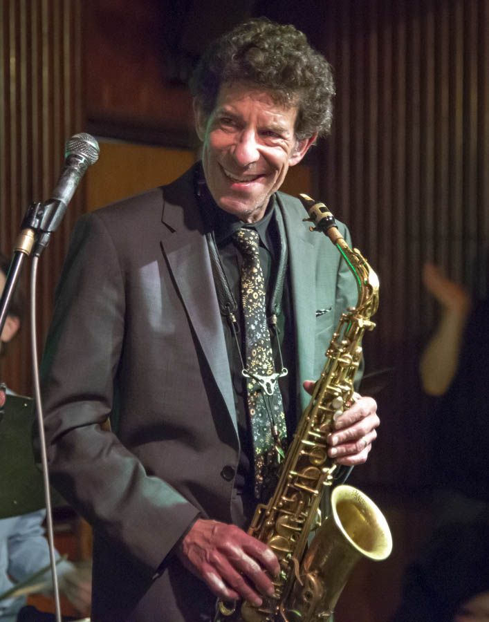 A saxophonist in a gray jacket and patterned tie smiling beside a microphone on a stage.
