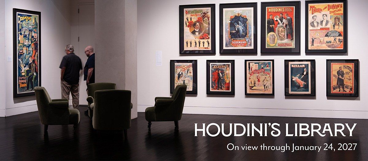 A gallery with framed vintage posters on a dark wall and two visitors looking at them, with a Houdini's Library caption.