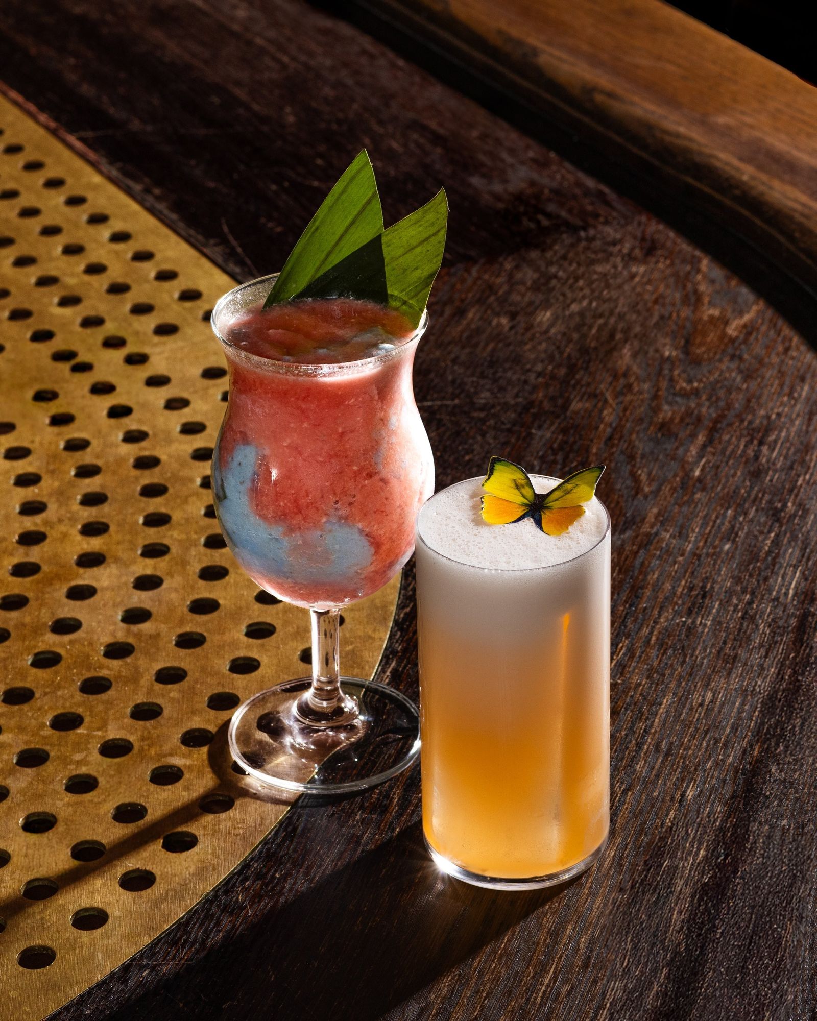 A pink frozen cocktail with a mint-leaf garnish and a pale cocktail with an orange peel, on a perforated metal tray.