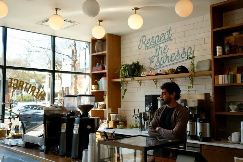A bright cafe counter with espresso machines and wall shelves under a Respect the Espresso sign, with large windows.