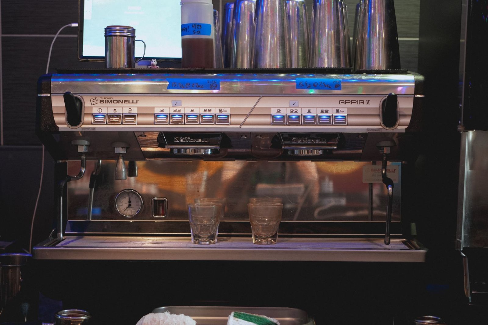 A silver espresso machine on a counter with a row of glass cups below it in a dim cafe.