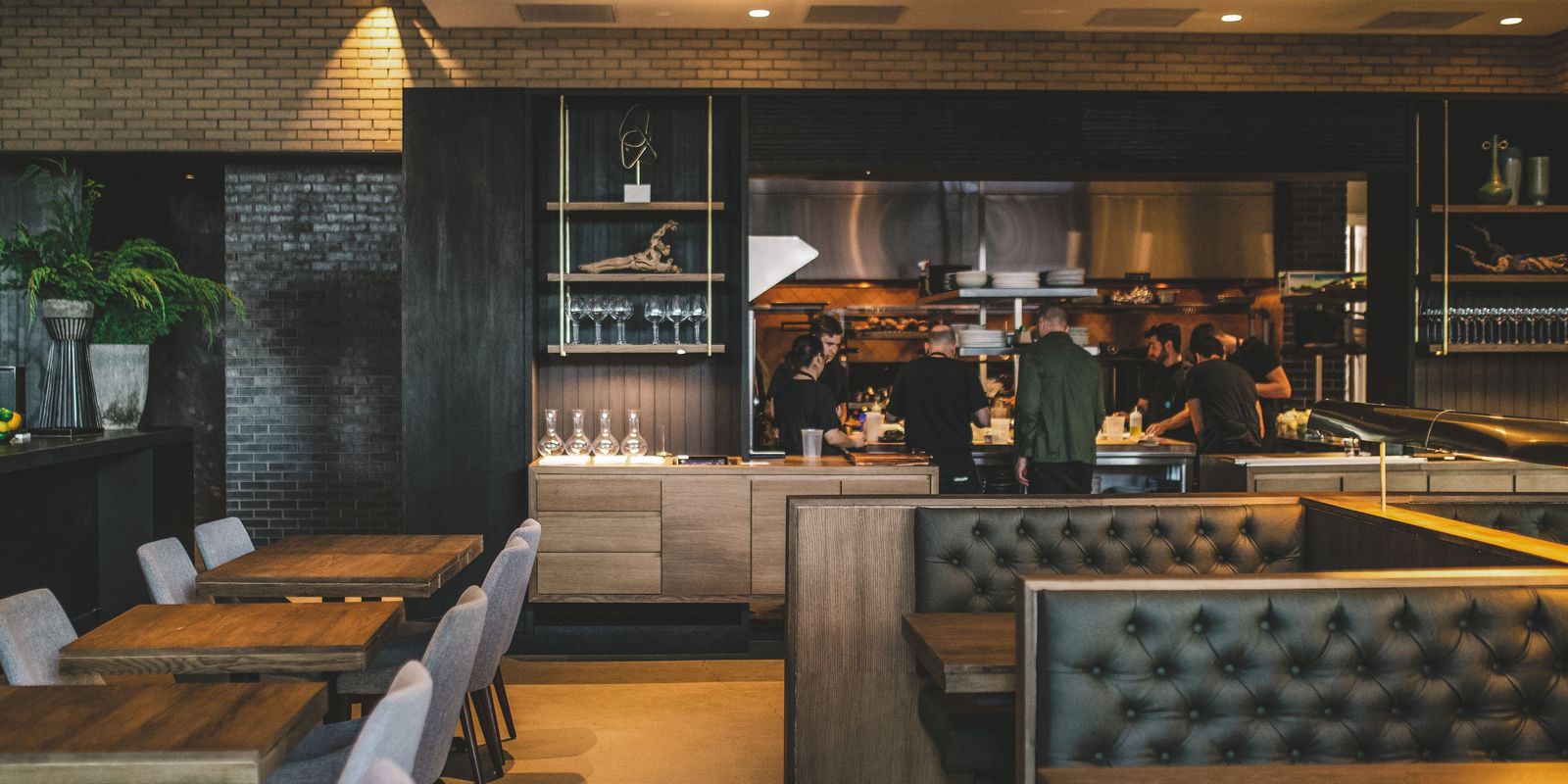 A dark dining room with an open kitchen, tufted leather banquettes and wooden tables.