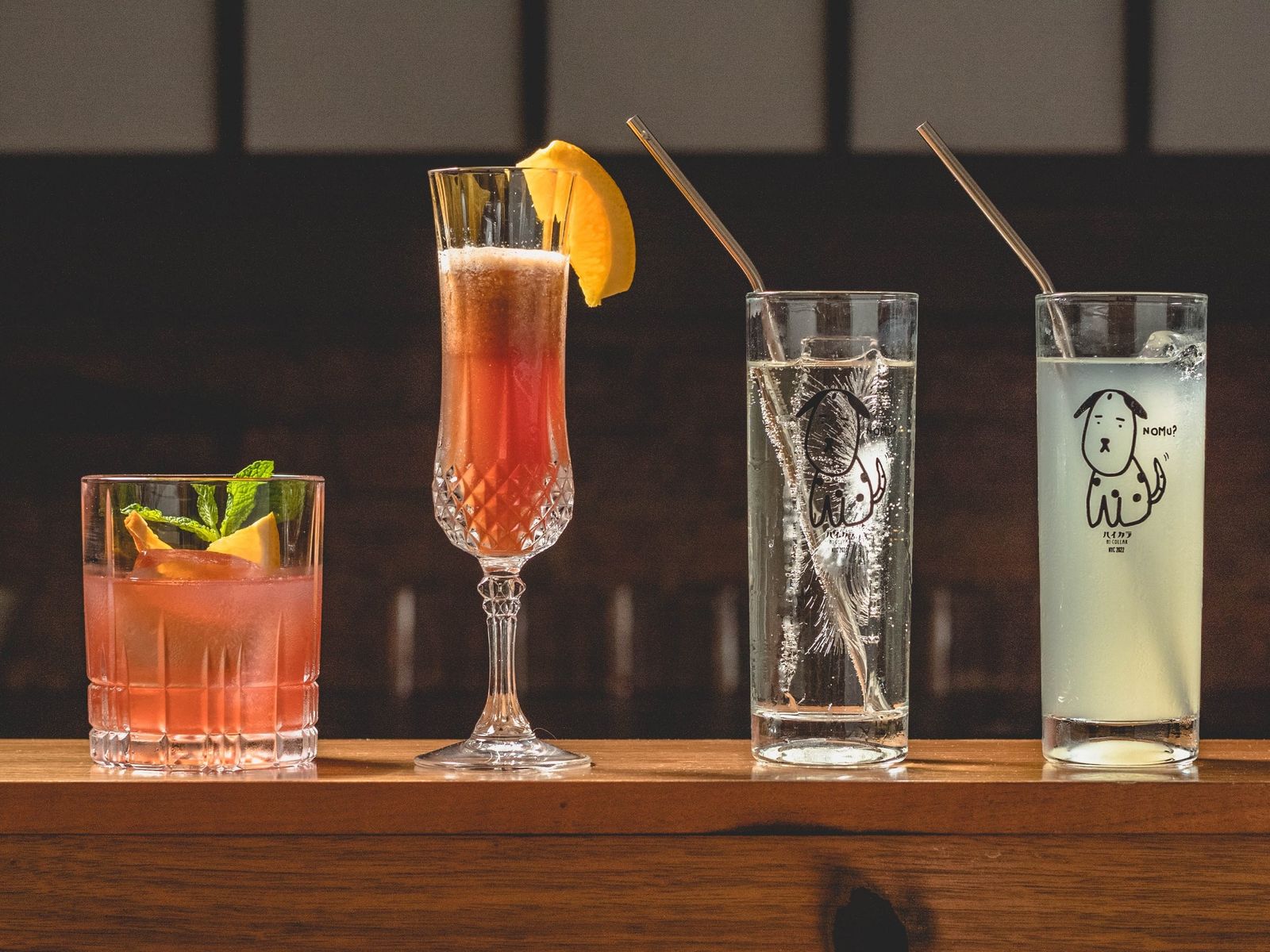A row of cocktails and tall glasses of lemonade on a wooden bar, with a window screen behind.