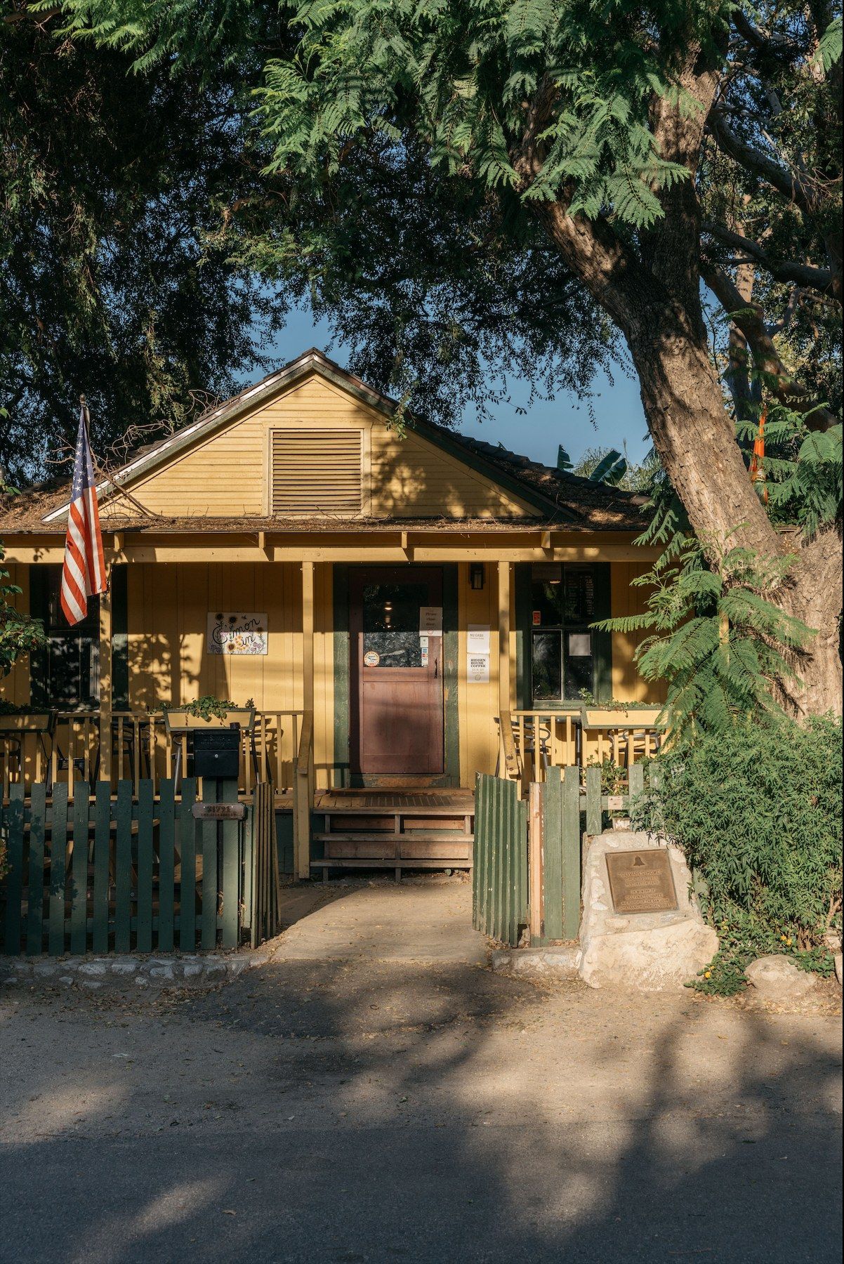 A small yellow wooden house with a porch, under large trees.