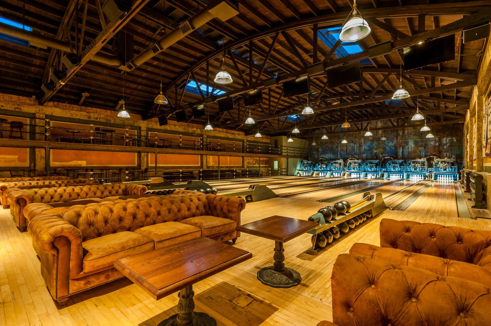 Bowling lanes under an exposed-beam ceiling, with tufted leather sofas and wooden tables in the foreground.
