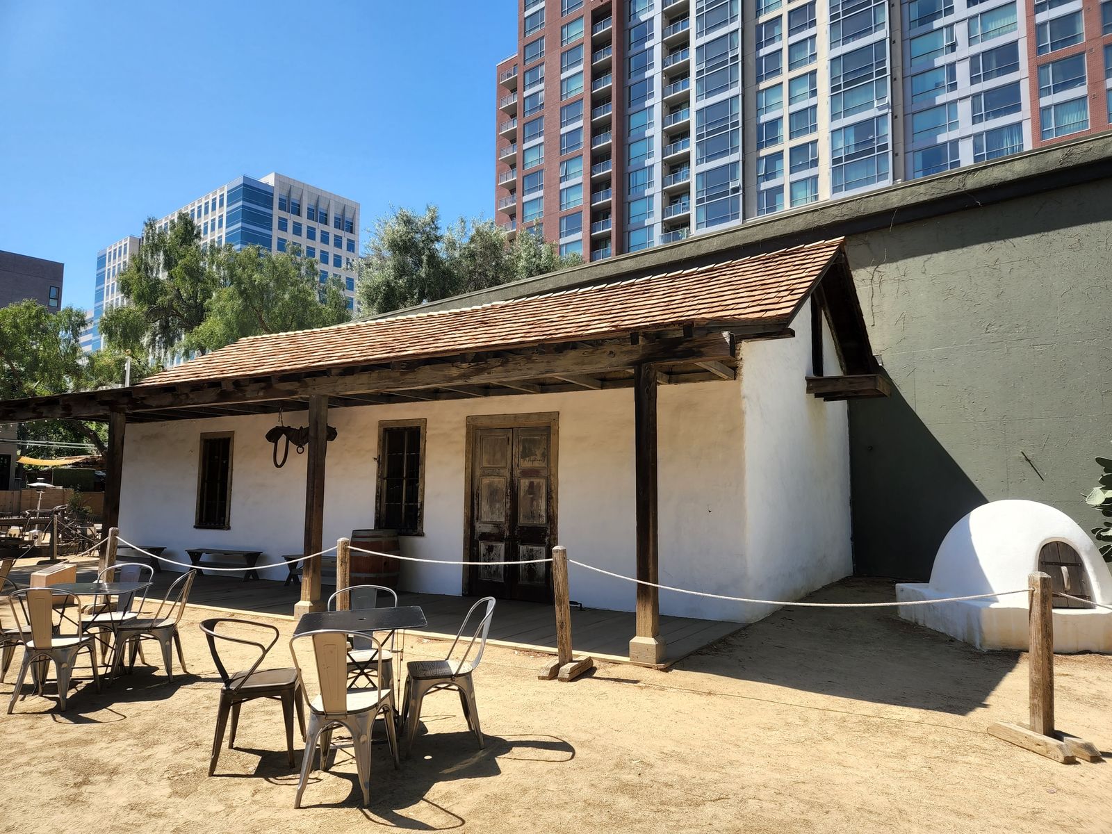 Low whitewashed adobe building with a tiled roof and outdoor tables, with high-rise buildings behind