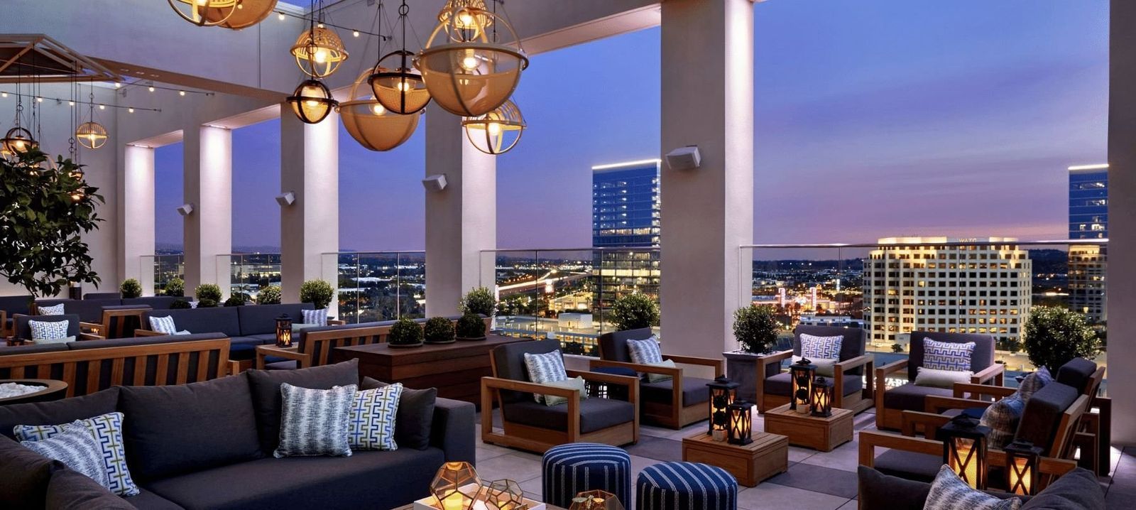 A rooftop lounge with sofas, pendant lanterns and city lights at dusk.