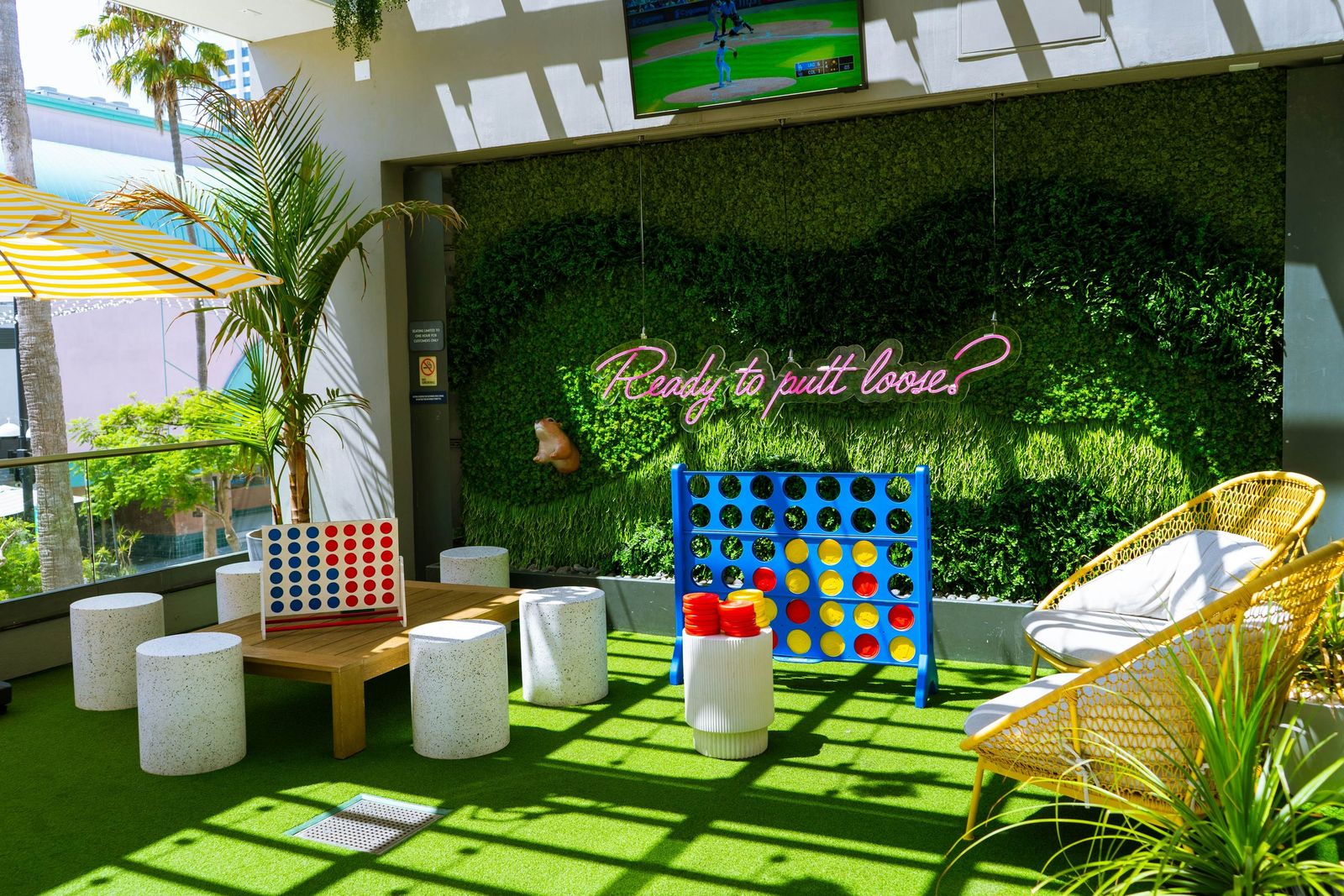 Mini golf lounge with a hedge wall and neon sign, giant Connect Four and white stools on artificial turf