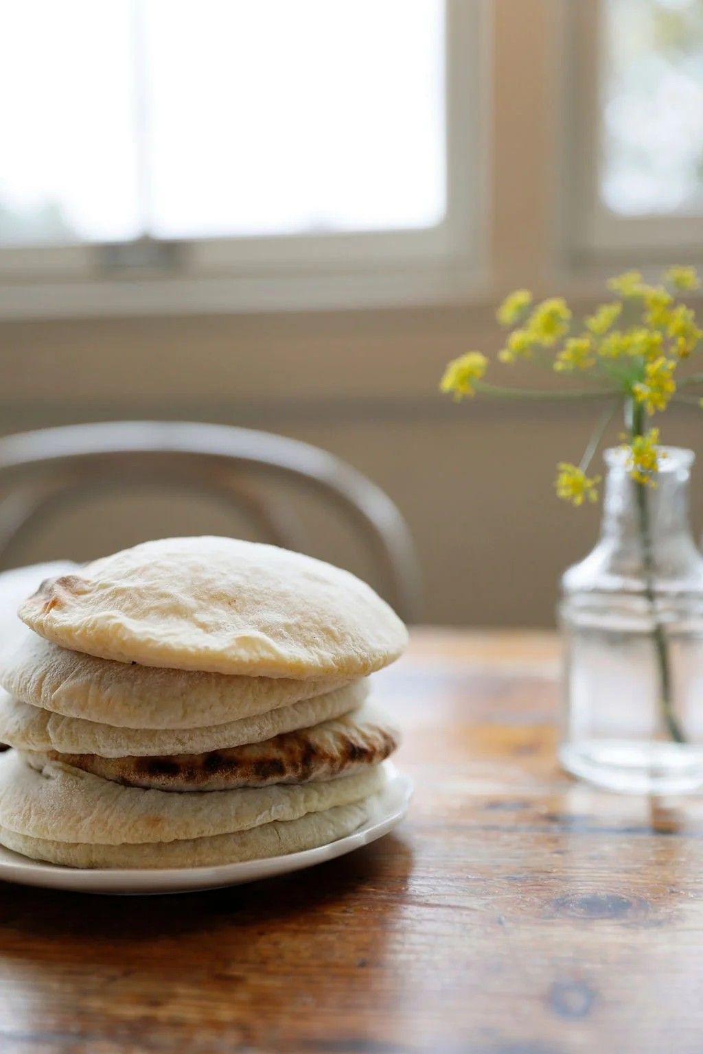 A stack of flatbreads on a plate at a window table with a small vase of yellow flowers.
