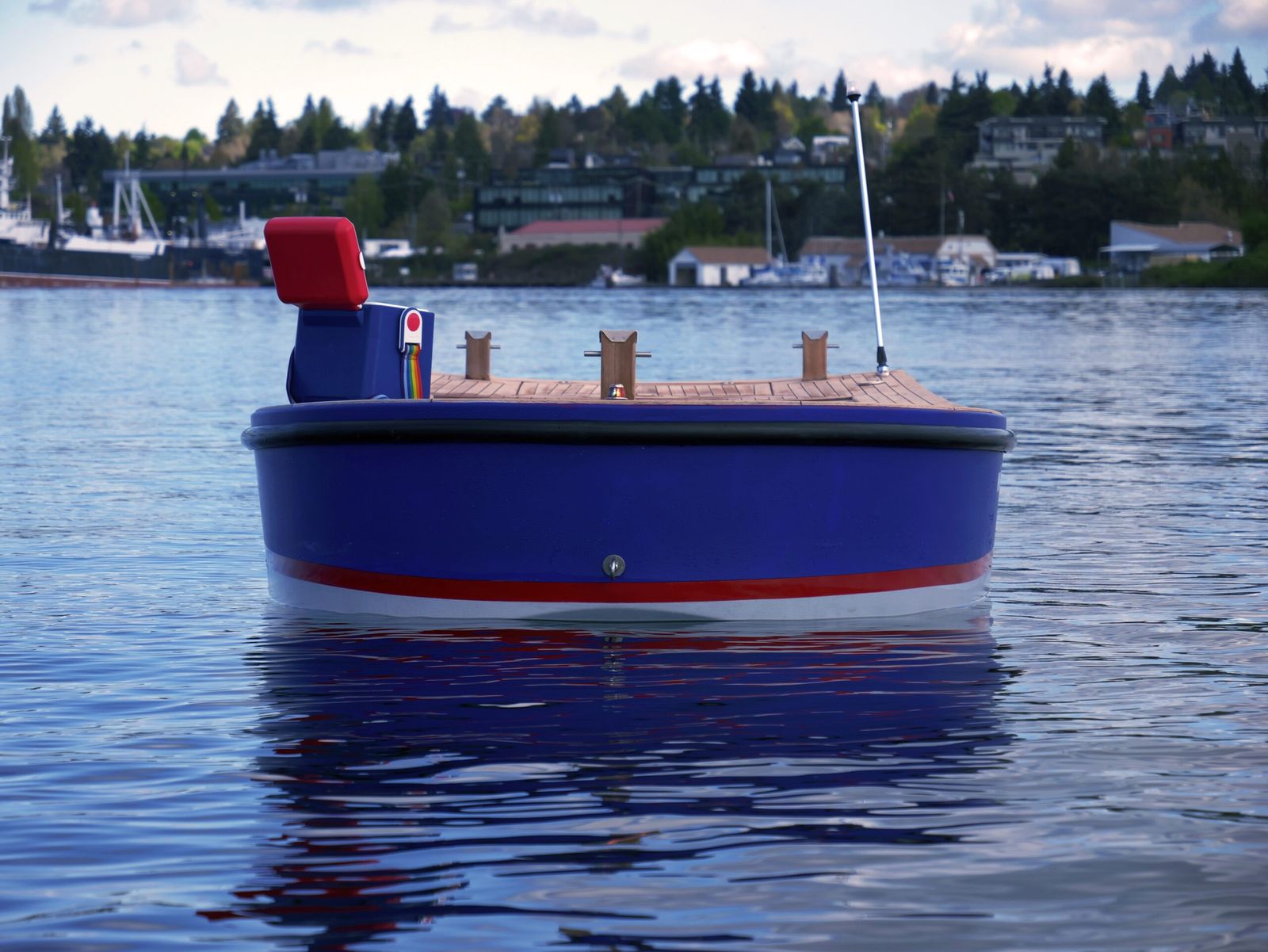 A small blue boat with a wooden deck floating on a lake, with a tree-lined shore behind it.