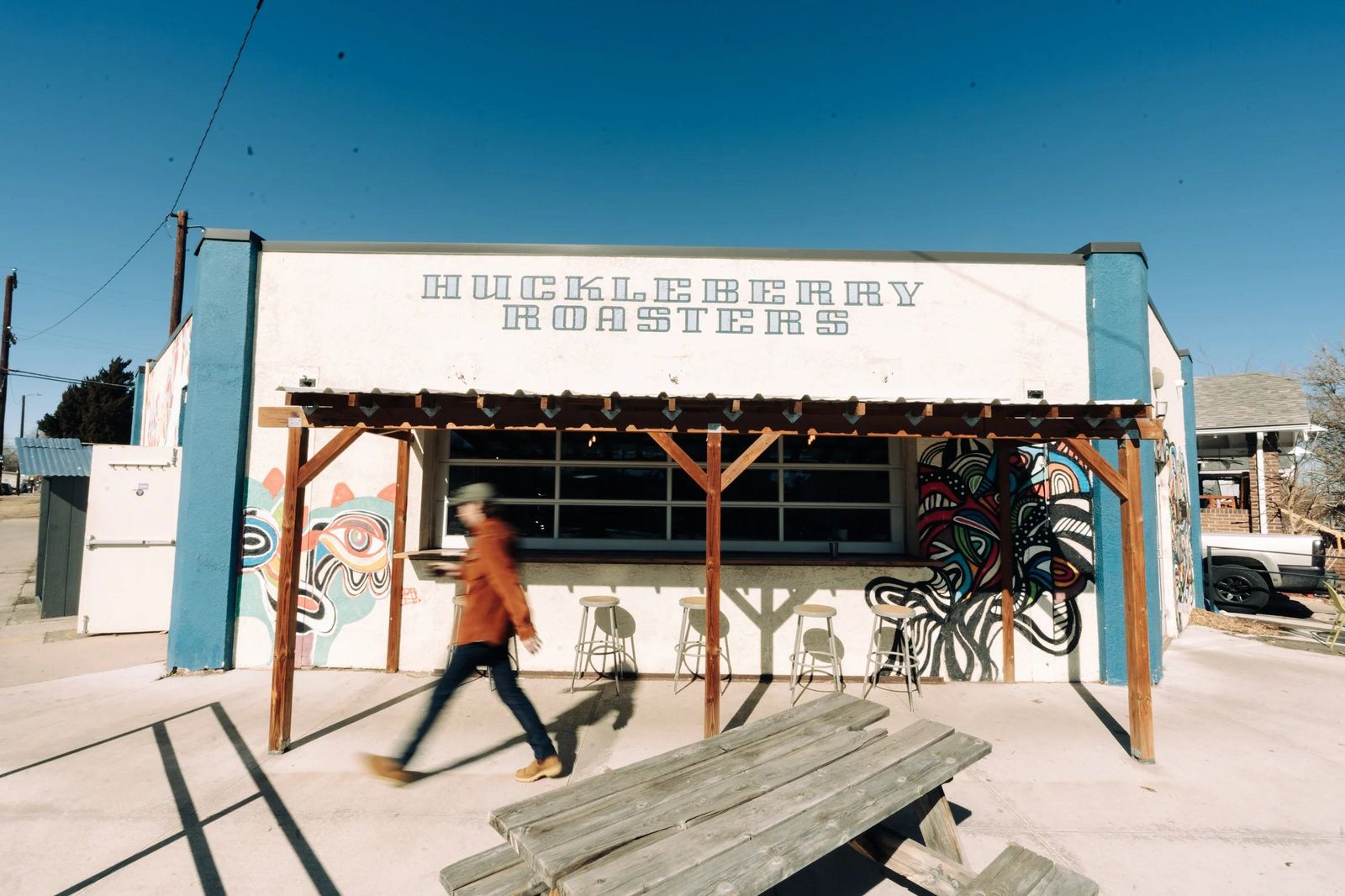 A white and blue cafe building with a covered patio and a person walking past a picnic table.